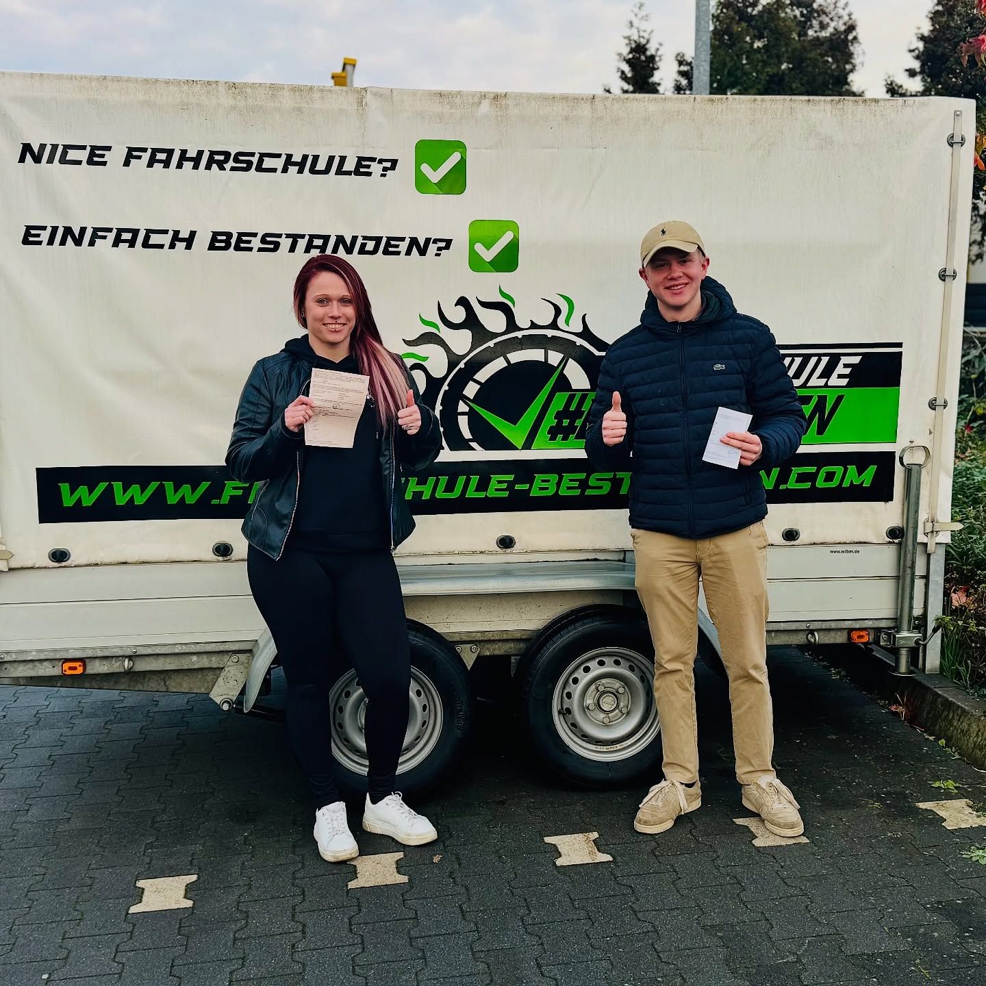 Zwei Fahrschüler mit ihren Prüfungsbescheinigungen vor dem Fahrschul-Anhänger „Nice Fahrschule? Einfach bestanden?“